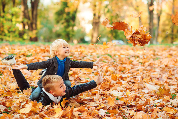 Χαριτωμένα παιδιά παίζουν και γελάνε στην φθινοπωρινή βόλτα. Ευτυχισμένοι αδελφοί ρίχνουν φύλλα του φθινοπώρου έξω. Διακοπές το φθινόπωρο. Μοντέρνα παιδιά στο πάρκο. Παιδική μόδα. Αγόρια που παίζουν με φύλλα. - Φωτογραφία, εικόνα