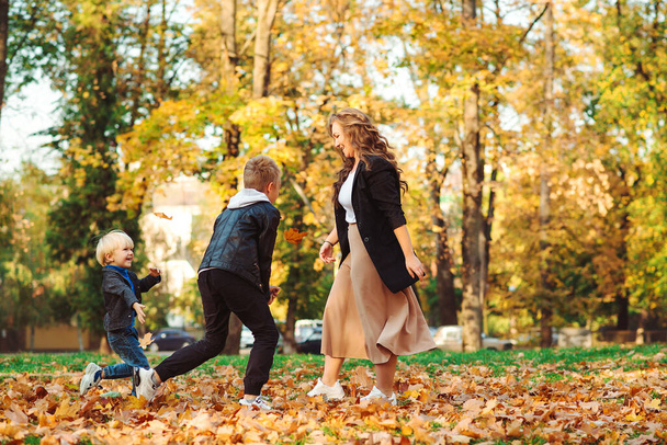 Anne ve çocuklar sonbahar yapraklarını parka atıyorlar. Sonbahar tatili. Anne ve oğulları dışarıda eğleniyorlar. Sonbahar doğasında şık bir aile yürüyüşü. Aile, moda ve yaşam tarzı - Fotoğraf, Görsel