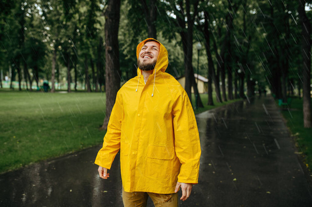 Mężczyzna w płaszczu przeciwdeszczowym cieszący się deszczem w letnim parku. Szczęśliwy mężczyzna w deszczu peleryna na ścieżce spacerowej, mokra pogoda w alejce - Zdjęcie, obraz