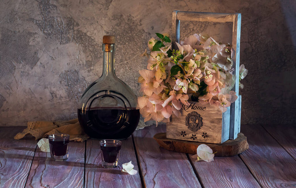 Nature morte avec liqueur maison dans une bouteille et des piles et un panier en bois avec des fleurs sur une table en bois close-up - Photo, image