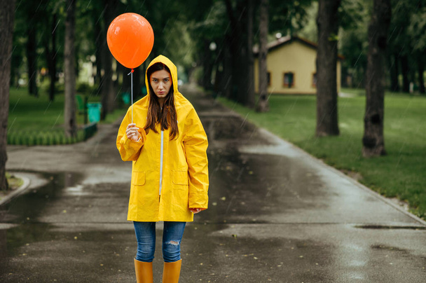 Θλιβερή γυναίκα με μπαλόνι που περπατάει στο πάρκο τις βροχερές μέρες. Γυναίκα με κάπα και λαστιχένιες μπότες, υγρός καιρός στο σοκάκι, μοναξιά - Φωτογραφία, εικόνα