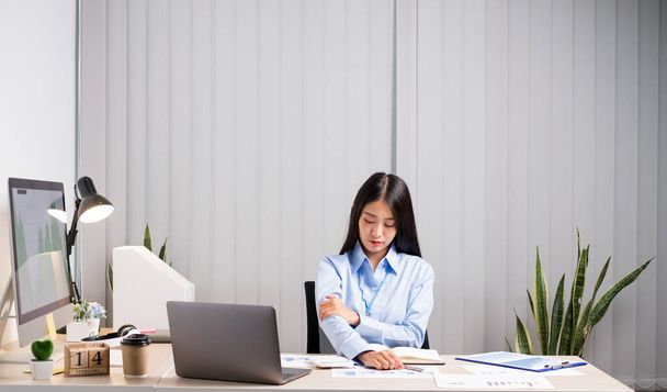 Aziatische vrouwelijke accountant is moe van het werken in een stoel, stretching, en masseren van haar armen voor ontspanning tijdens het werken hard op kantoor. - Foto, afbeelding