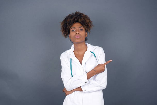 Excited surprised African American doctor woman wearing medical uniform with trendy hairstyle points happily aside on copy space of gray background for your fashion clothing advertising information or promotional text. - Photo, Image
