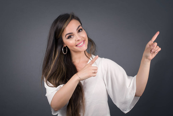 Werbekonzept. Positive, hübsche, kaukasische Frau mit strahlendem Lächeln im Jeanshemd auf grauem Hintergrund zeigt mit ihren beiden Zeigefingern auf leeren Kopierraum. - Foto, Bild