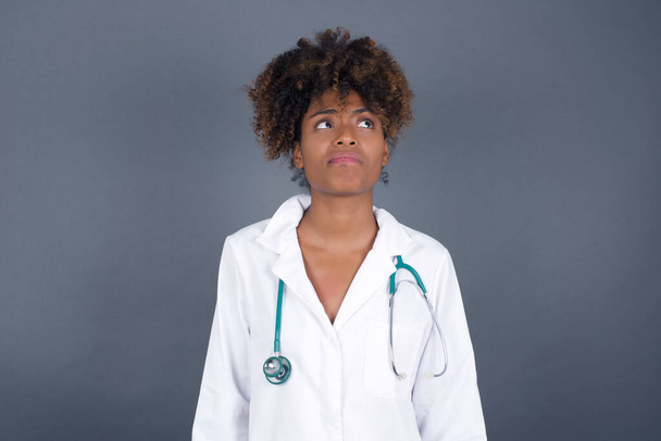 Troublesome puzzled  African American doctor female wearing medical uniform  frowns face, bites lip, raises eyebrows, looks up, has problems, dressed medical uniform, isolated over gray background, has regret look. - Фото, изображение