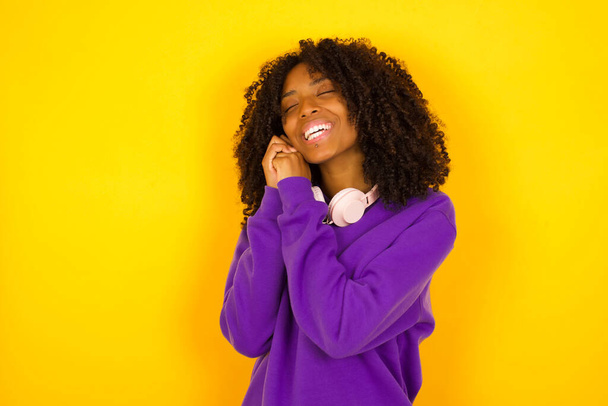 Young beautiful African American woman wearing purple knitted sweater pleased  against yellow  background.   - Foto, afbeelding