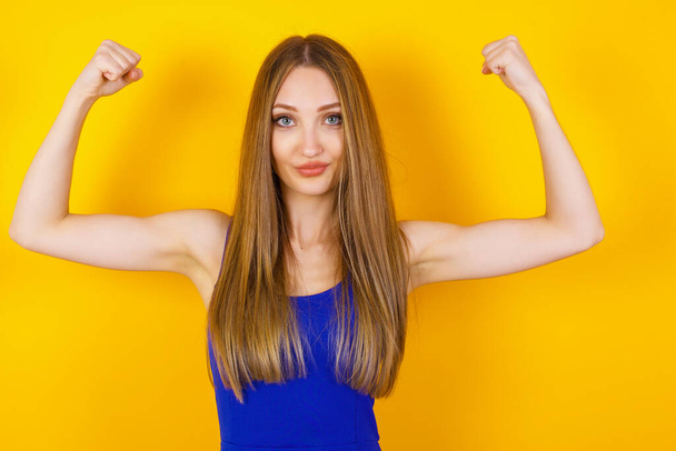 Waist up shot of caucasian woman raises arms to show her muscles feels confident in victory, looks strong and independent, smiles positively at camera, stands against studio background. Sport concept. - Photo, Image
