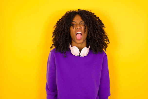 Молодая красивая афроамериканка в фиолетовом трикотажном свитере показывает язык на жёлтом фоне.   - Фото, изображение