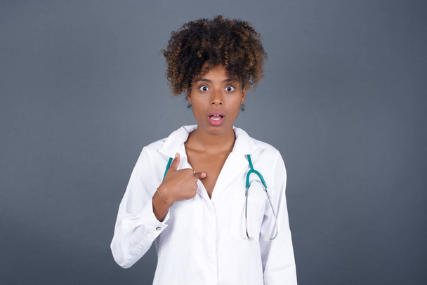 Meglepett afro-amerikai doktornő, aki orvosi egyenruhát visel, és döbbent arckifejezéssel mutogatott magára: Szürke háttérrel elszigetelve. - Fotó, kép
