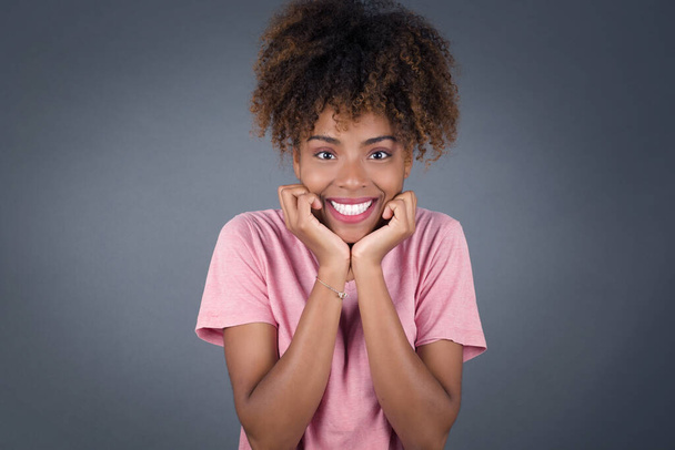 Мрійлива молода красива афроамериканка тримає руки притиснуті під підборіддям, виглядає з щасливим виразом, має зубну посмішку, ізольовані на сірому фоні - Фото, зображення