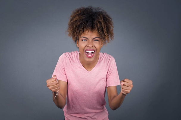 Portrait of beautiful african american woman looks with excitement at camera, keeps hands raised over head, notices something unexpected, isolated over gray wall. - Photo, Image