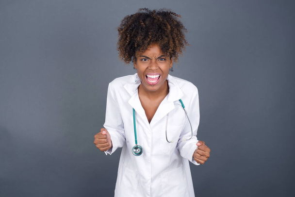 Портрет афроамериканської жінки-лікаря в медичній формі виглядає з хвилюванням на камеру, тримає руки підняті над головою, помічає щось несподіване, ізольоване над сірою стіною. Прекрасна жінка реагує на раптові новини. - Фото, зображення