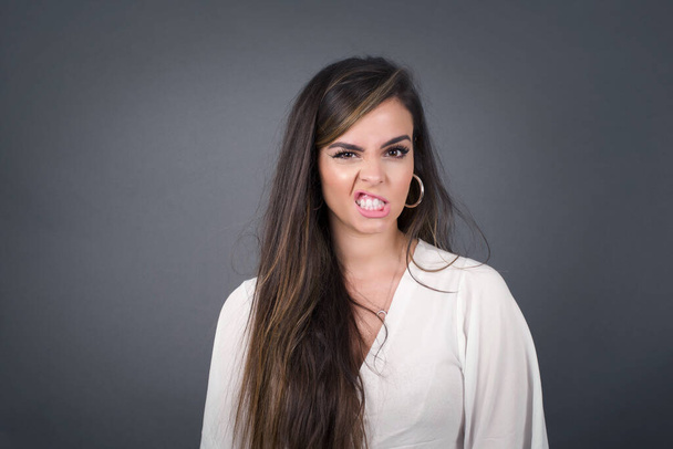 Mad crazy woman clenches teeth angrily, being annoyed with coming noise, dressed in fashionable clothes, isolated over gray background. Negative feeling concept. - Foto, imagen