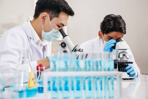 Двое ученых-мужчин носят маску для лица, работая в лаборатории, проверяя раствор с помощью микроскопа - Фото, изображение