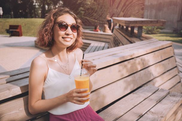 Happy woman with slim figure drinks freshly squeezed orange juice from a plastic cup. Useful and harmful beverages - 写真・画像