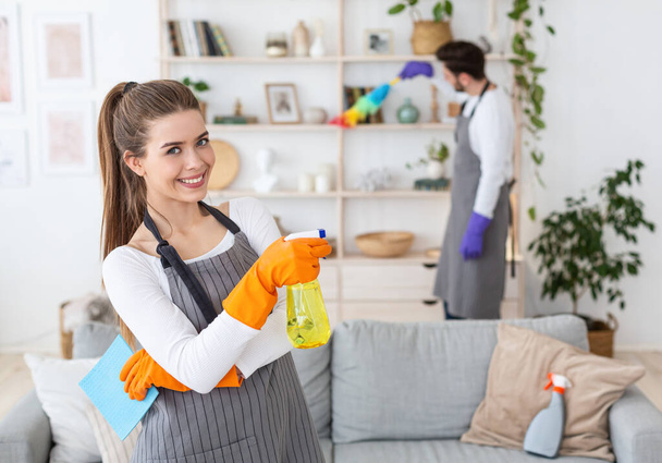 Уборка дома вместе. Улыбающаяся женщина с брызгами вблизи, указывает на занятого мужа - Фото, изображение
