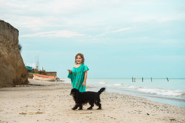 Child walking alone with dog at the beach. Kid in a big t-shirt with little black dog. Barefoot on sand. Ocean. - Photo, Image