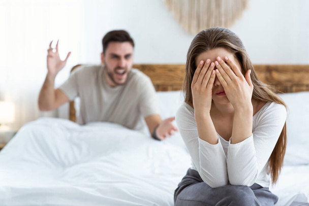 Triste esposa cubre su cara con las manos y se sienta en la cama, marido jura y gestos en el interior del dormitorio - Foto, imagen
