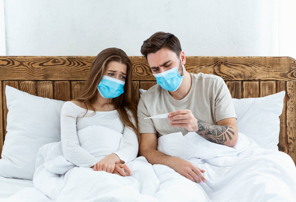 Quarantine together. Sad young man and woman in protective masks look at thermometer - Fotó, kép