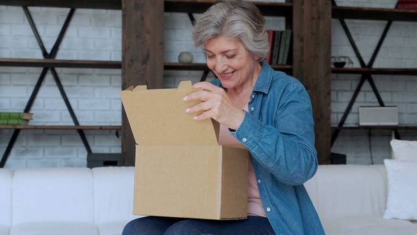 Elderly woman sit on couch in living room open cardboard box shopping online at home. Delivery concept - Photo, Image
