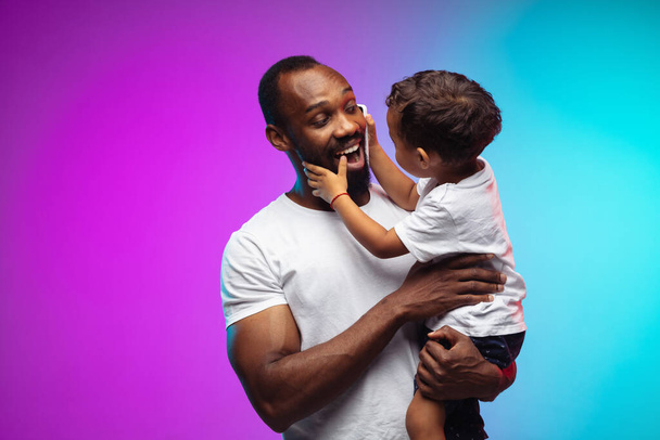 African-american father and son portrait on gradient studio background in neon - Φωτογραφία, εικόνα
