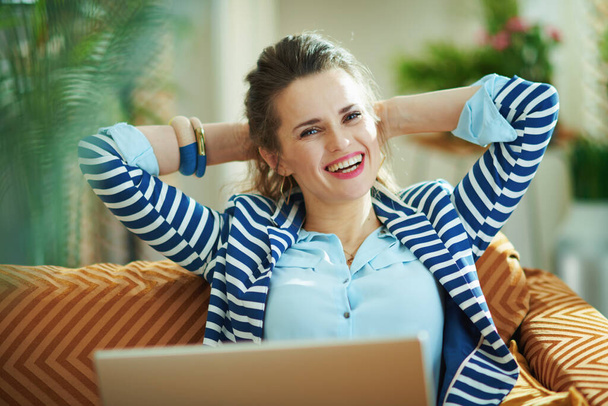 smiling middle age woman in blue blouse and striped jacket sitting on couch with laptop in the modern house in sunny day. - Foto, Imagem