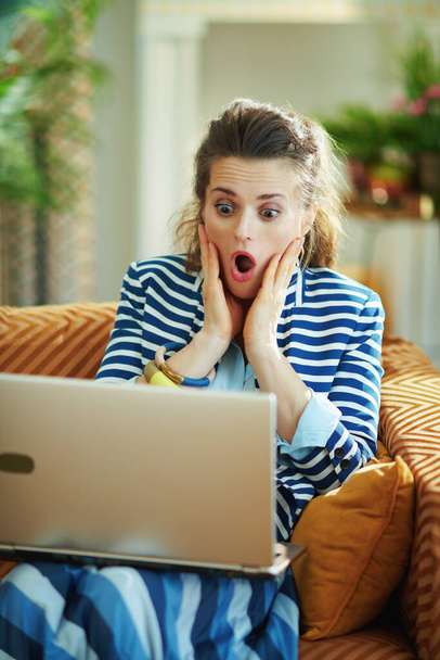 shocked modern female in blue blouse and striped jacket sitting on couch with laptop in the modern house in sunny day. - Zdjęcie, obraz