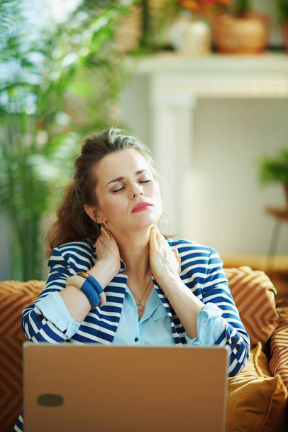 stressed 40 years old housewife in blue blouse and striped jacket having neck pain sitting on couch with laptop in the modern house in sunny day. - Foto, immagini