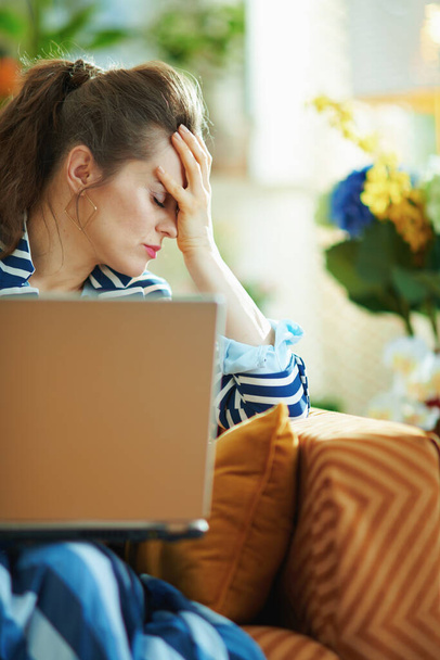 stressed young housewife in blue blouse and striped jacket sitting on sofa with laptop at modern home in sunny day. - Foto, Bild