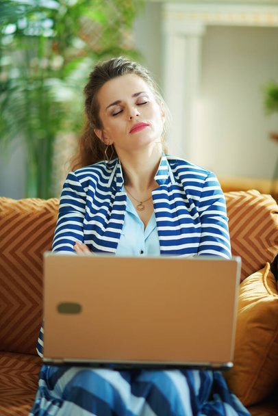 stressed modern 40 years old housewife in blue blouse and striped jacket sitting on divan with laptop in the modern house in sunny day. - Φωτογραφία, εικόνα