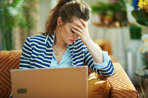 stressed elegant middle age woman in blue blouse and striped jacket sitting on couch with laptop in the modern living room in sunny day. - Fotó, kép