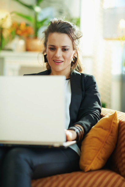 surprised stylish woman in white blouse and black jacket in the modern living room in sunny day reading text on a laptop while sitting on couch. - Zdjęcie, obraz