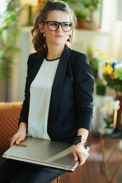 trendy 40 years old woman in white blouse and black jacket at modern home in sunny day in glasses holding closed laptop. - Фото, изображение