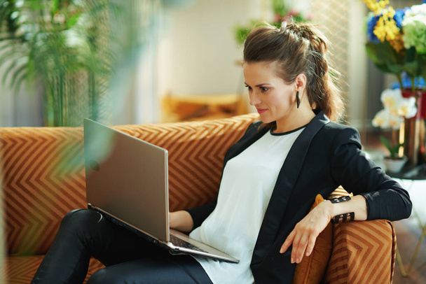 stylish 40 years old housewife in white blouse and black jacket in the modern living room in sunny day writing on a laptop while sitting on sofa. - Фото, изображение