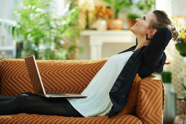 relaxed trendy 40 years old woman in white blouse and black jacket with laptop sitting on sofa in the modern living room in sunny day. - 写真・画像