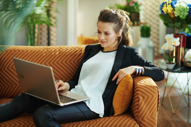 elegant middle age housewife in white blouse and black jacket at modern home in sunny day surfing web on a laptop while sitting on divan. - Foto, Bild