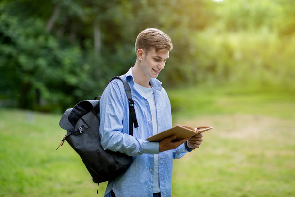 Концепция обучения. Красавчик-студент читает книгу на открытом воздухе, отдыхает в кампусе - Фото, изображение