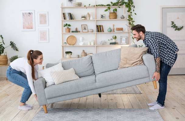 Moving or permutation furniture at home. Happy guy and girl raise sofa in interior of living room - Photo, Image