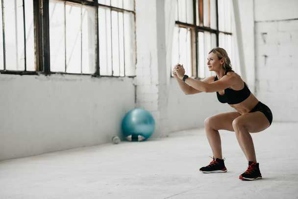 Femme mince sportive en vêtements de sport avec tracker de fitness pratiquant des exercices de squat en studio à la lumière du jour - Photo, image
