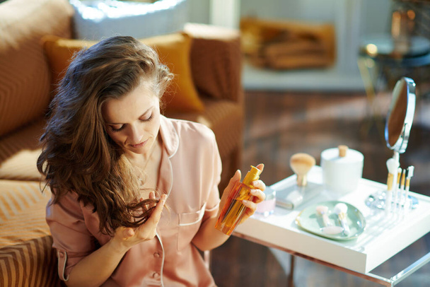 concerned middle age housewife in pajamas with long brunette wavy hair holding hair oil bottle and checking hair ends near table with toiletries in the modern house in sunny winter day. - 写真・画像