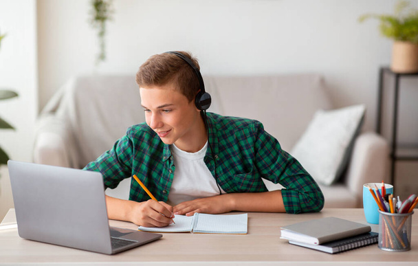 Joyful teen guy writing at notebook and looking at laptop - Valokuva, kuva