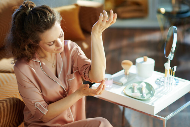 elegant 40 years old housewife in pajamas applying moisturiser on elbow near table with toiletries in the modern living room in sunny winter day. - Foto, imagen