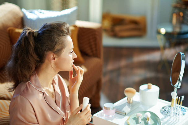 modern 40 years old woman in pajamas with white cosmetic tube applying lip contour cream near table with toiletries in the modern house in sunny winter day. - 写真・画像