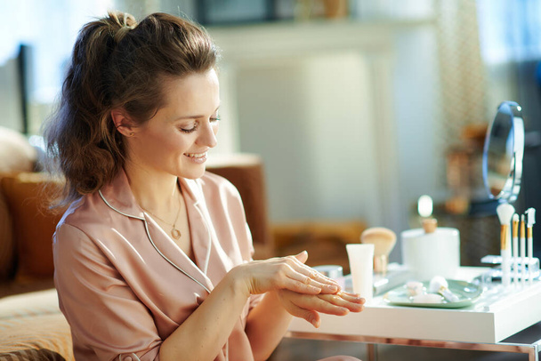 smiling stylish woman in pajamas applying hand cream near table with toiletries at modern home in sunny winter day. - Foto, immagini