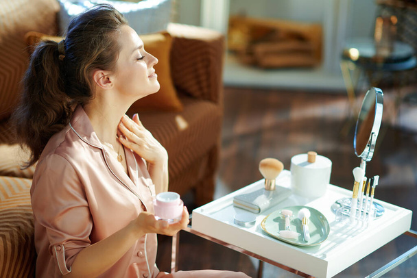 happy modern woman in pajamas with pink cosmetic jar applying neck cream and looking in mirror near table with toiletries at modern home in sunny winter day. - 写真・画像