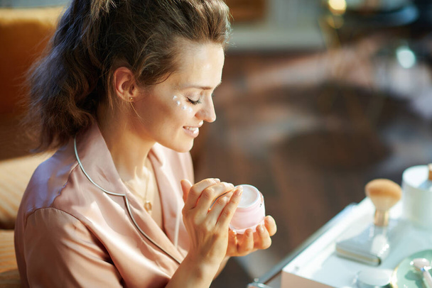happy elegant housewife in pajamas with pink cosmetic jar applying eye cream in the modern living room in sunny winter day. - Foto, Bild