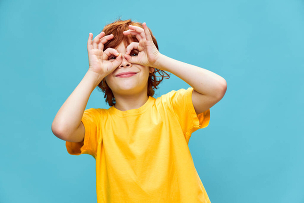 Niño feliz en una camiseta amarilla sostiene los dedos unidos delante de sus ojos  - Foto, imagen