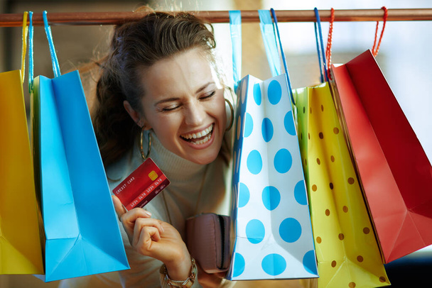 cheerful modern woman in white sweater and skirt peeking out from colorful shopping bags hanging on copper clothes rail with red credit card and purse. - Foto, Imagen