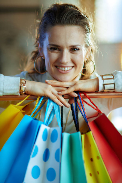 Portrait of happy trendy 40 years old woman in white sweater and skirt near colorful shopping bags hanging on copper clothes rail. - Foto, imagen