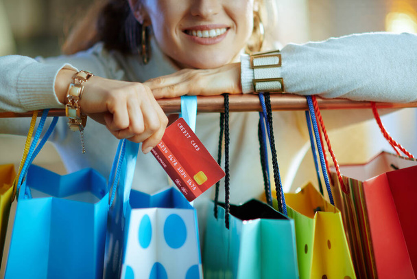 Closeup on smiling stylish 40 years old woman in white sweater and skirt with credit card near colorful shopping bags hanging on copper clothes rail. - Valokuva, kuva